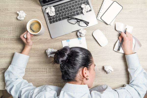 A woman at her messy desk. She has her head on the desk, arms splayed out and is clutching a cup of coffee. It is not a clean system!